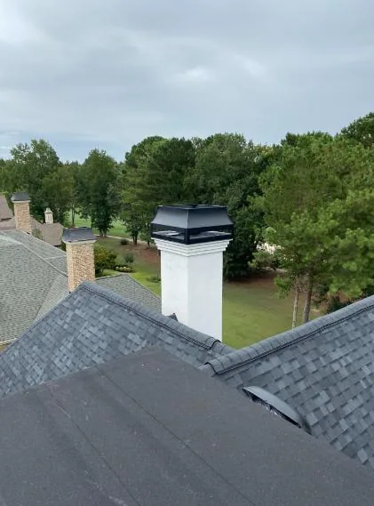 A finished white chimney with a black decorative shroud cap on a rooftop overlooking a lawn